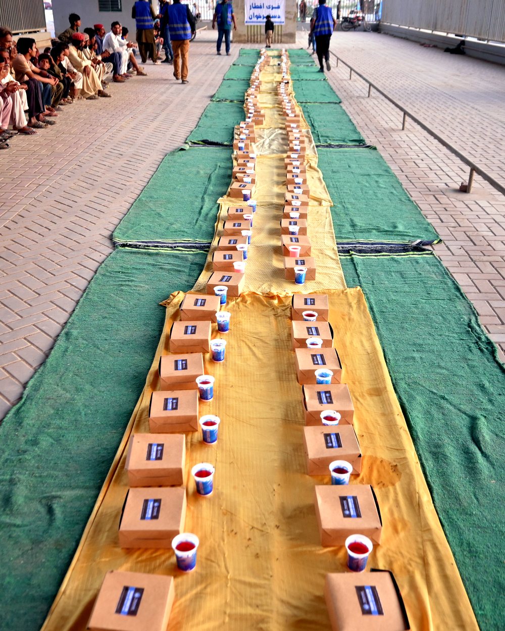 Long rows of iftar meal boxes and juice cups laid out on mats at a community iftar