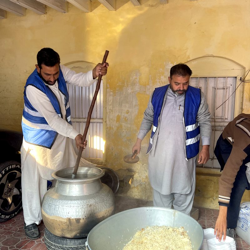 Two volunteers in blue vests cooking rice in a giant pot beside stacked meal boxes