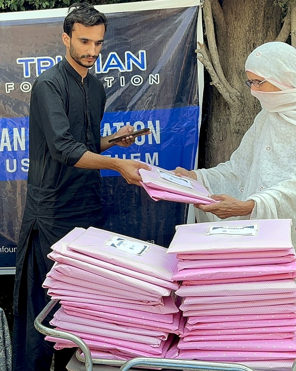 A volunteer hands an Eid gift parcel to an elderly woman beside a cart stacked with parcels